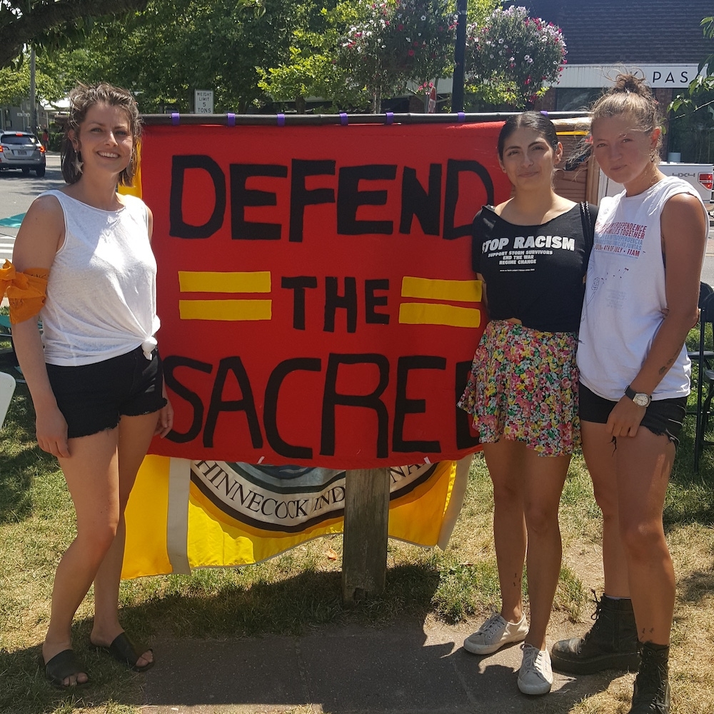 three young women stand besides red sign that reads defend the sacred